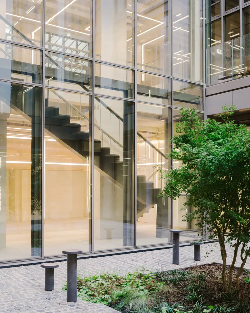 Glass curtain wall revealing floating staircase inside, with planted courtyard and bollards in foreground