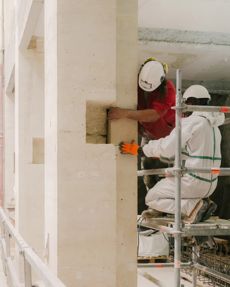 Two workers on scaffolding applying material to a concrete column corner joint