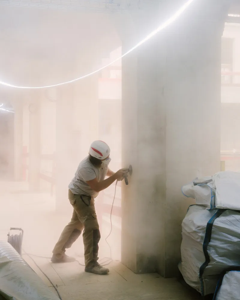 Worker sanding a cylindrical column in a dust-filled interior with curved cove lighting