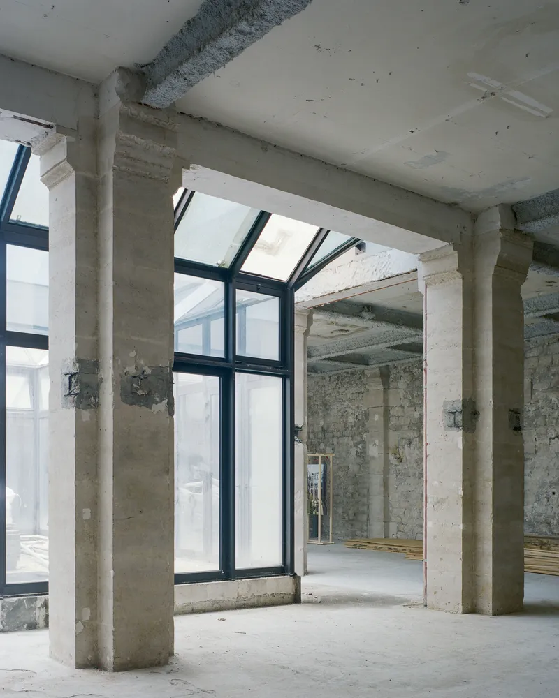 View through concrete columns toward a black-framed glazed opening in the existing structure