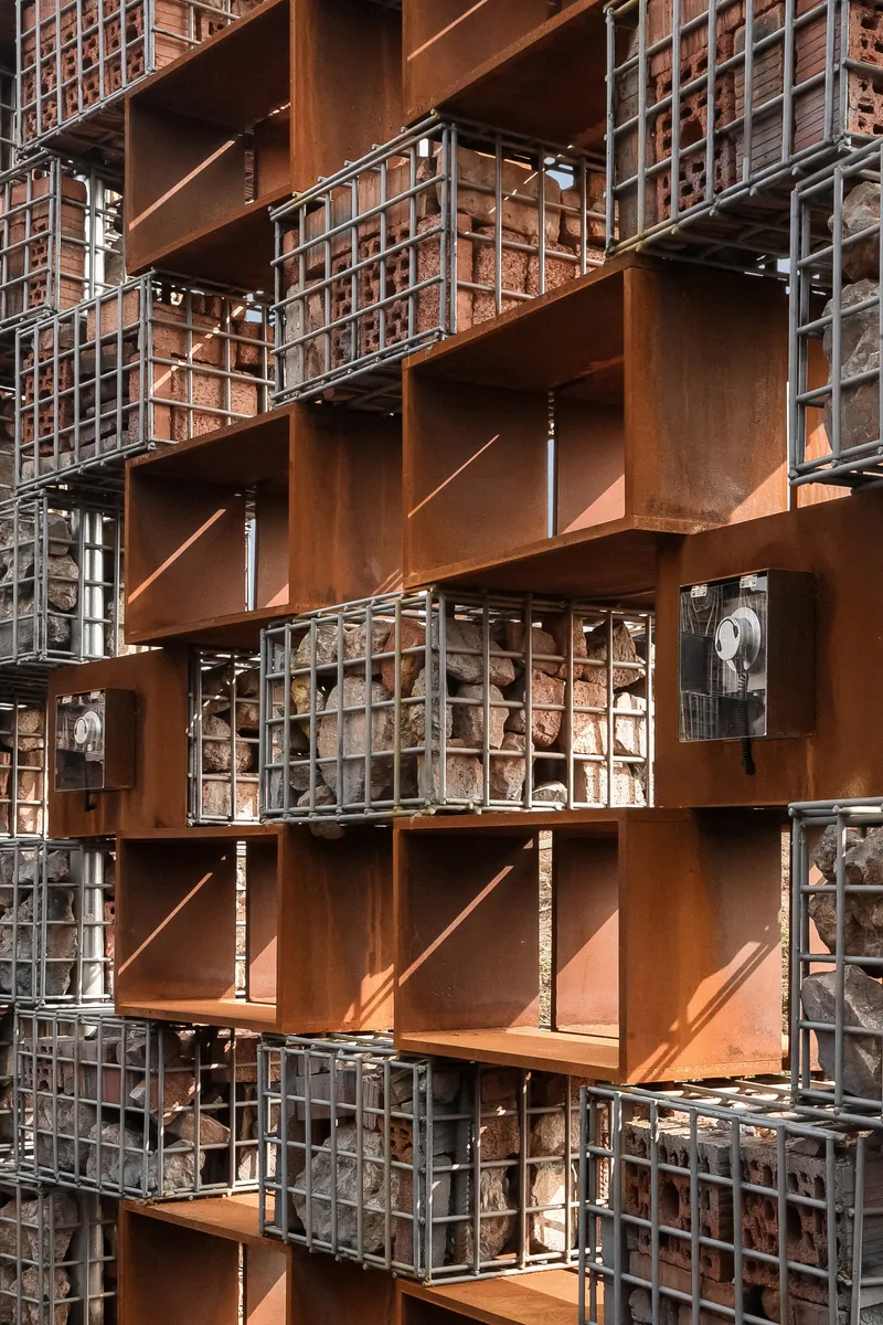 Close-up of the layered facade showing corten steel box frames alternating with wire mesh gabion panels filled with stone