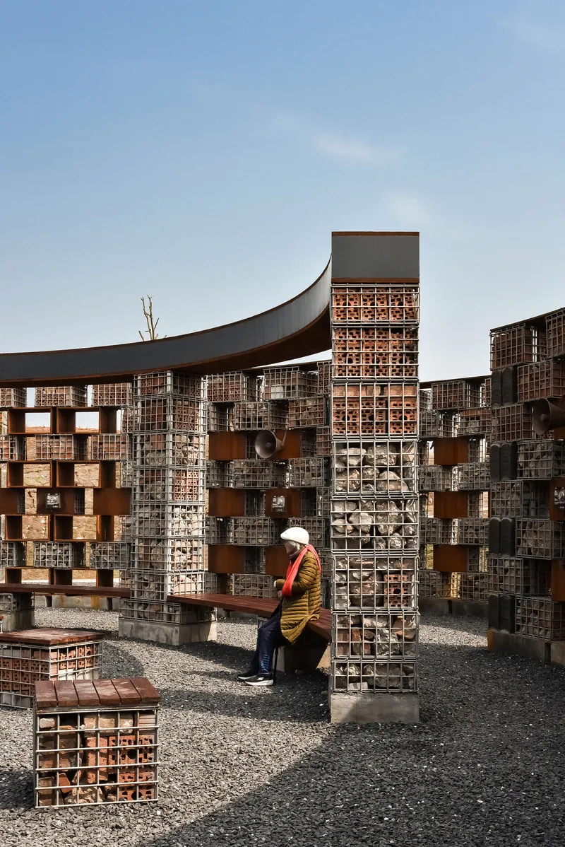 Seated visitor beside a brick column beneath the sweeping canopy and perforated brick facade with bare tree branch