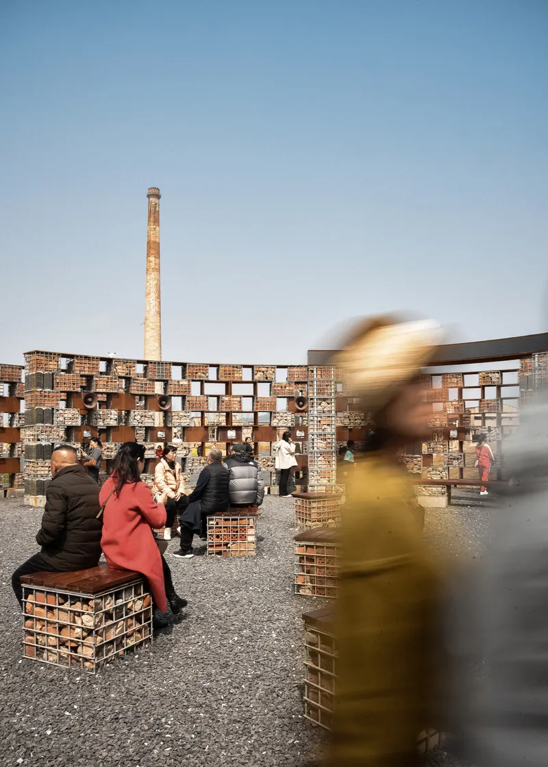 Gravel plaza at dusk with motion-blurred visitors on brick seating cubes and the brick chimney in the background