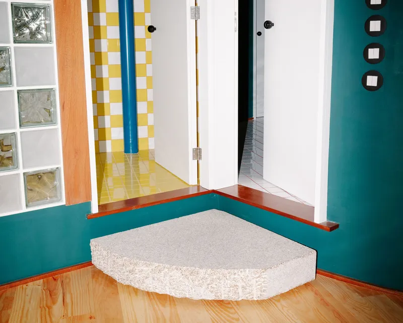 Corner detail of a white coral-textured plinth against teal walls and timber baseboard with glass block window