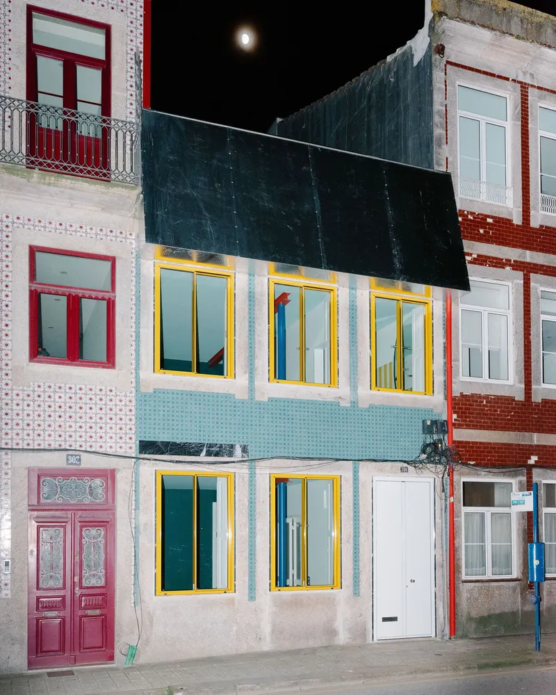 Street facade with yellow-trimmed windows, teal panels, and dark corrugated upper volume at night