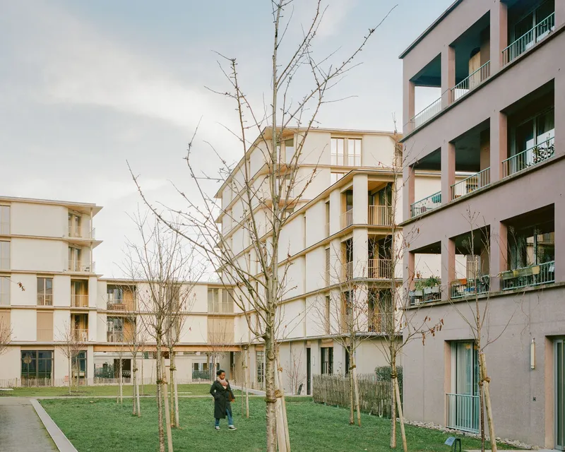 Shared courtyard with young bare trees and a pedestrian among three staggered housing blocks