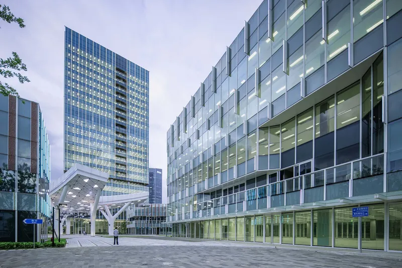 Pedestrian plaza between glass curtain wall towers and a cantilevered volume at dusk
