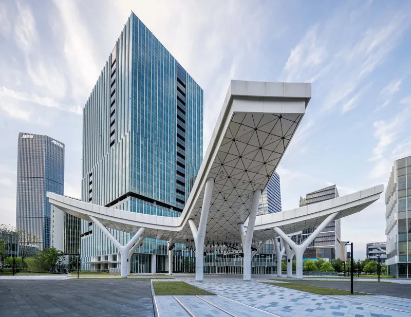 White triangulated canopy with branching columns sheltering the plaza beneath glass tower blocks under wispy clouds