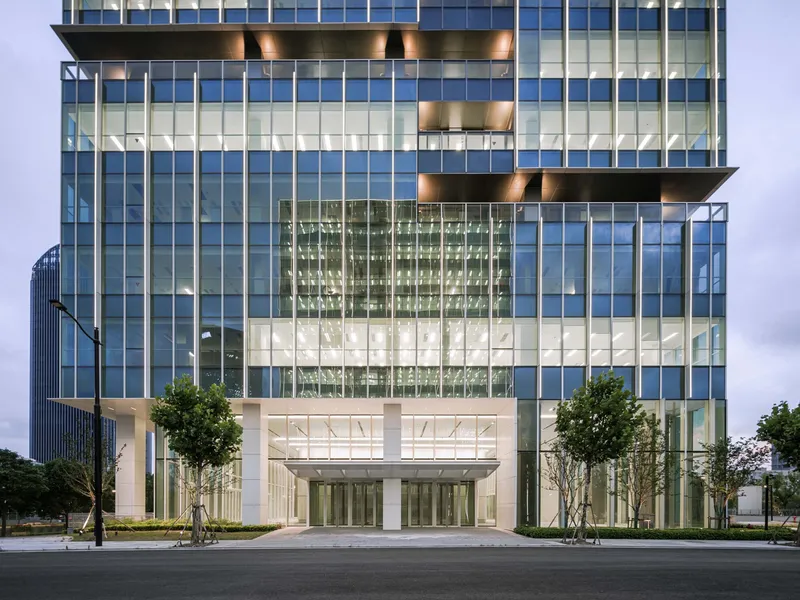 Tower entrance with glass lobby volume flanked by young trees and cantilevered floors above at twilight