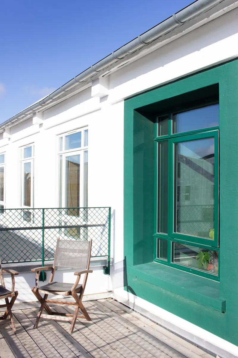 White rendered facade with green timber-framed window and folding chairs on the rooftop terrace under blue sky
