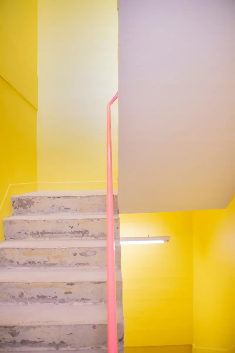 Concrete steps with pink handrail rising through yellow and white painted stairwell walls