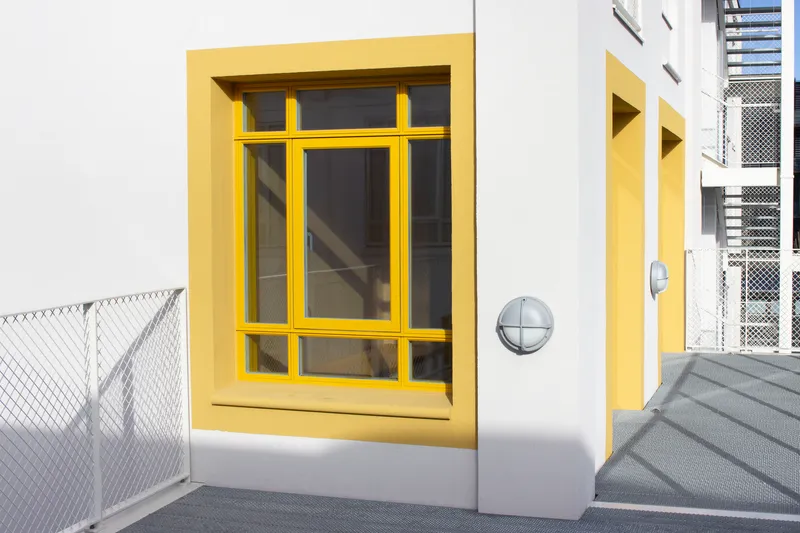 White facade with recessed yellow window frame and metal railing in afternoon light