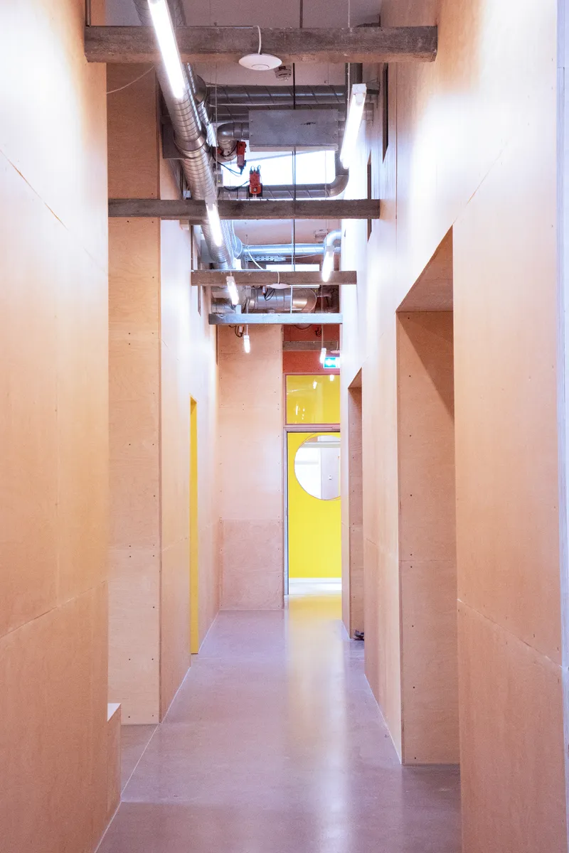Narrow corridor with plywood walls and exposed ceiling ductwork leading to a yellow doorway