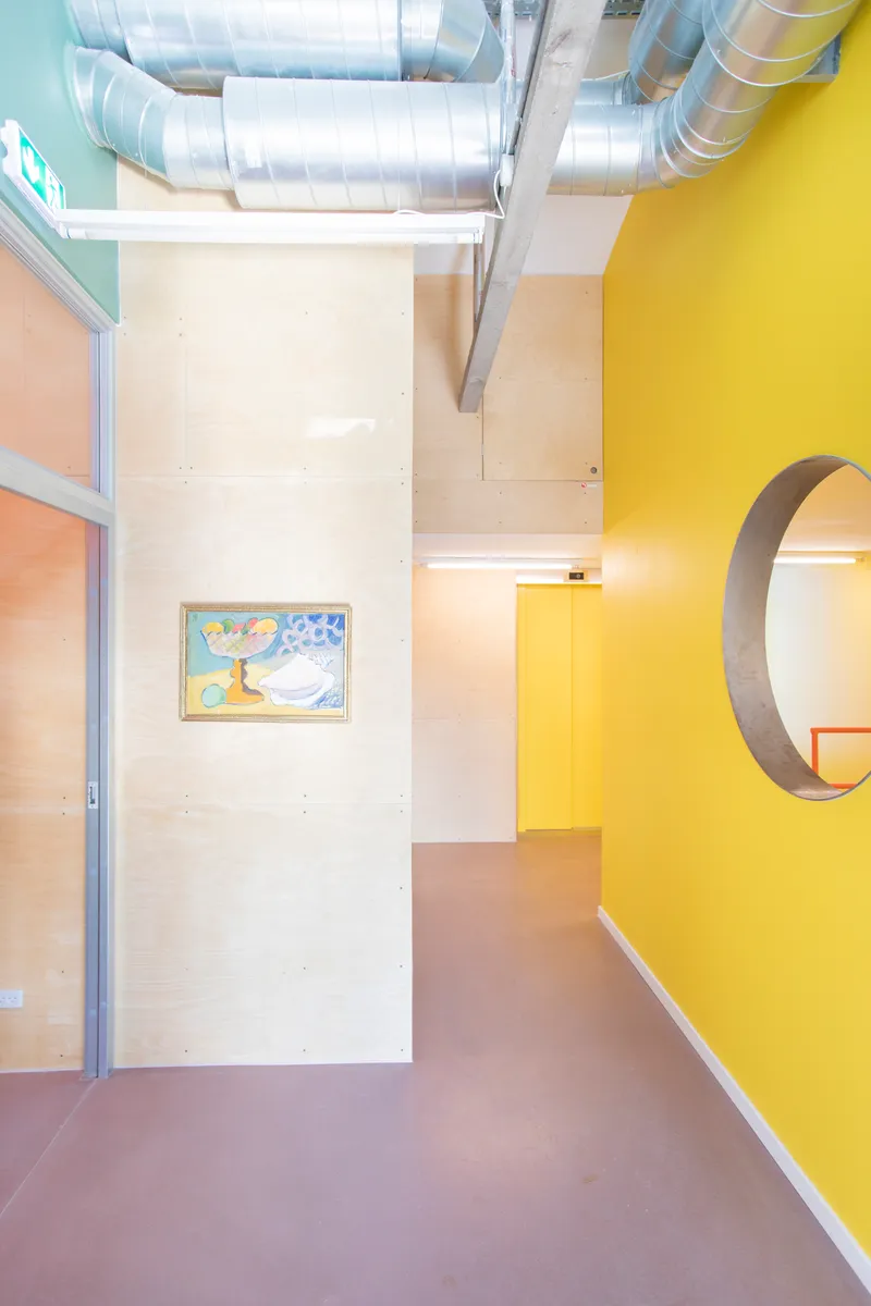 Interior corridor with exposed spiral ductwork overhead and a yellow accent wall with circular portal