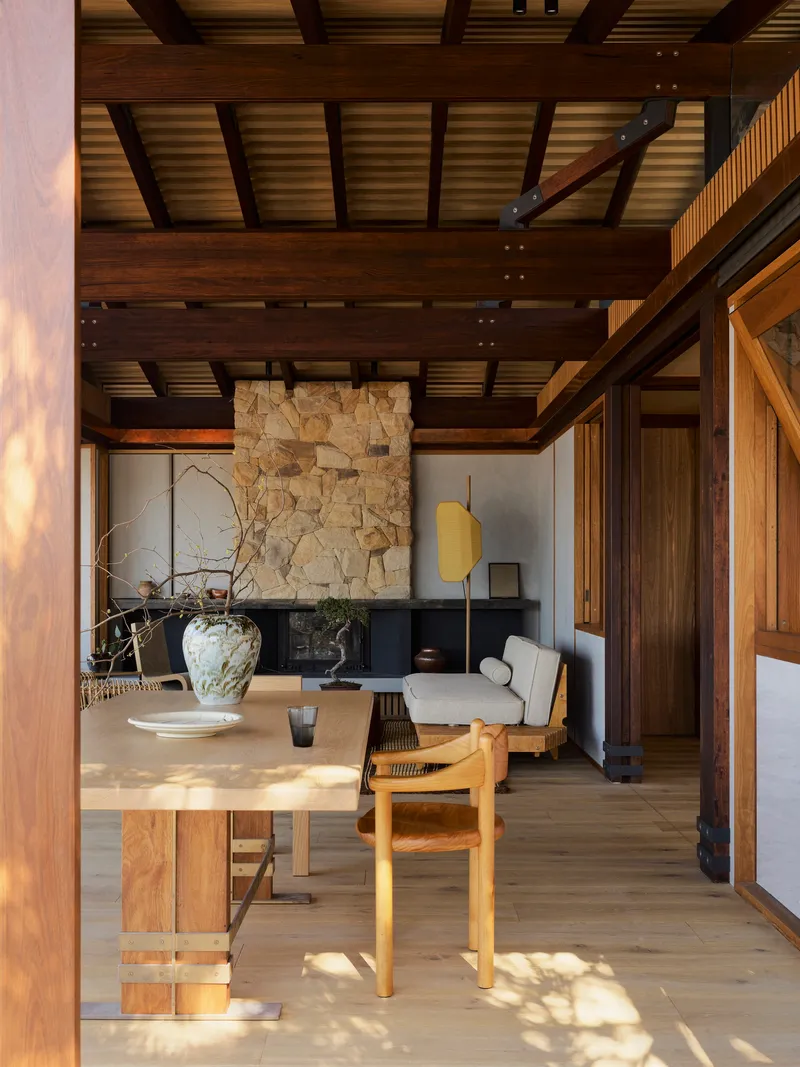 Open plan living space with exposed timber beams, stone fireplace wall and dappled afternoon sunlight