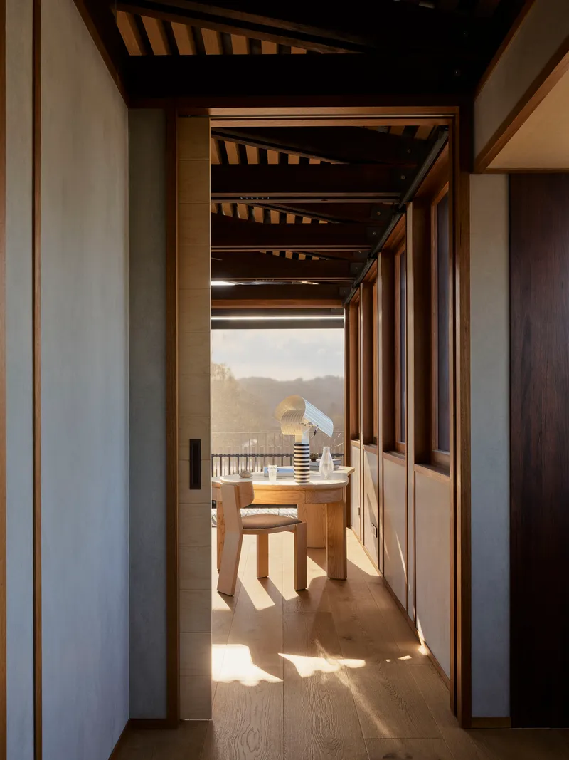 Narrow hallway with exposed timber ceiling beams leading to a desk and chair framed by sunlight