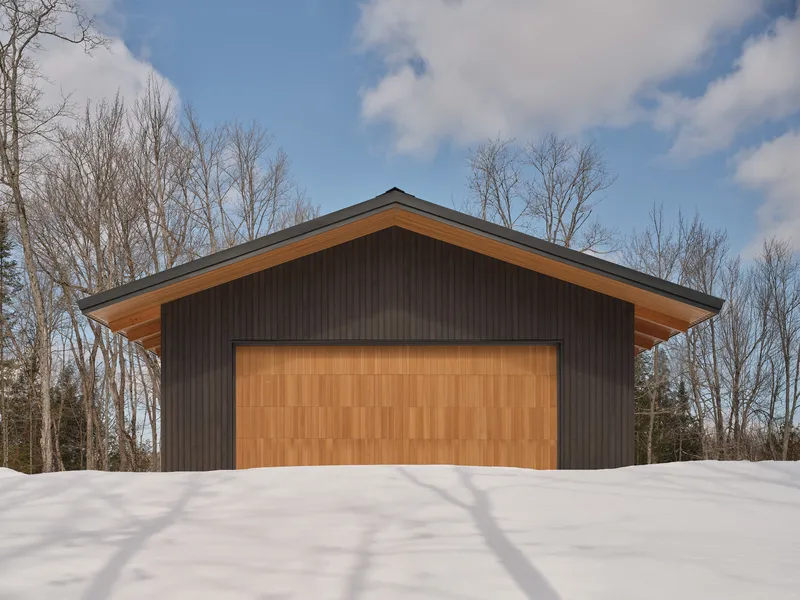 Front elevation showing timber garage door and extended roof overhangs casting shadows on fresh snow