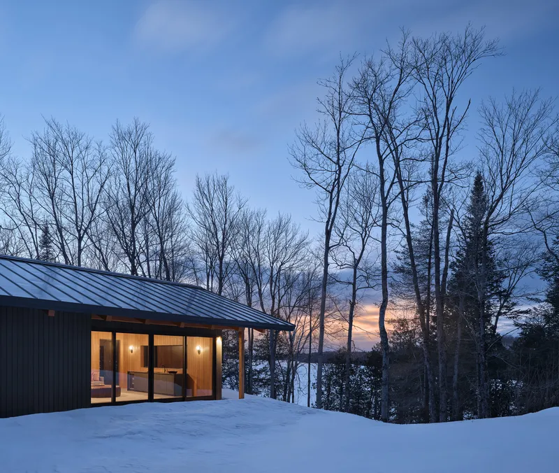 Glass-walled pavilion with metal roof and overhanging eave overlooking bare trees and a frozen lake at dusk