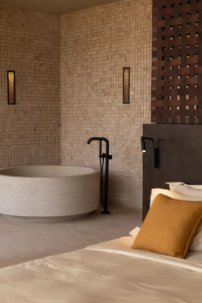 Circular stone bathtub with freestanding black tap against a small-tile mosaic wall in low light
