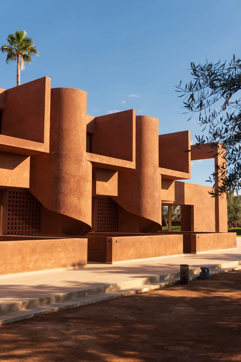 Terracotta facade with cylindrical columns and lattice screens beside an olive tree in afternoon sunlight