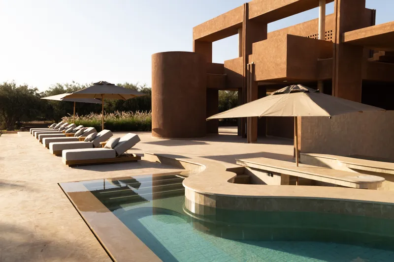 Curved pool edge with rows of white loungers beneath the sculptural terracotta facade at dusk