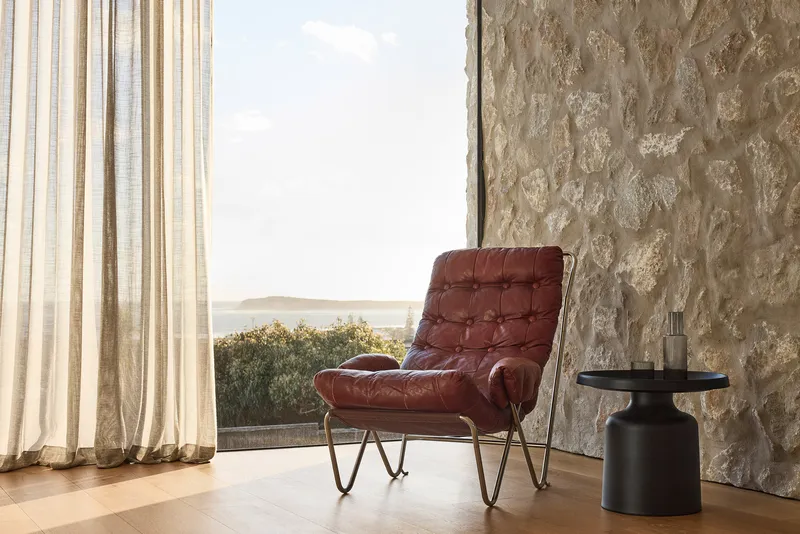 Interior corner with textured stone wall, sheer curtains, and leather armchair overlooking a coastal view