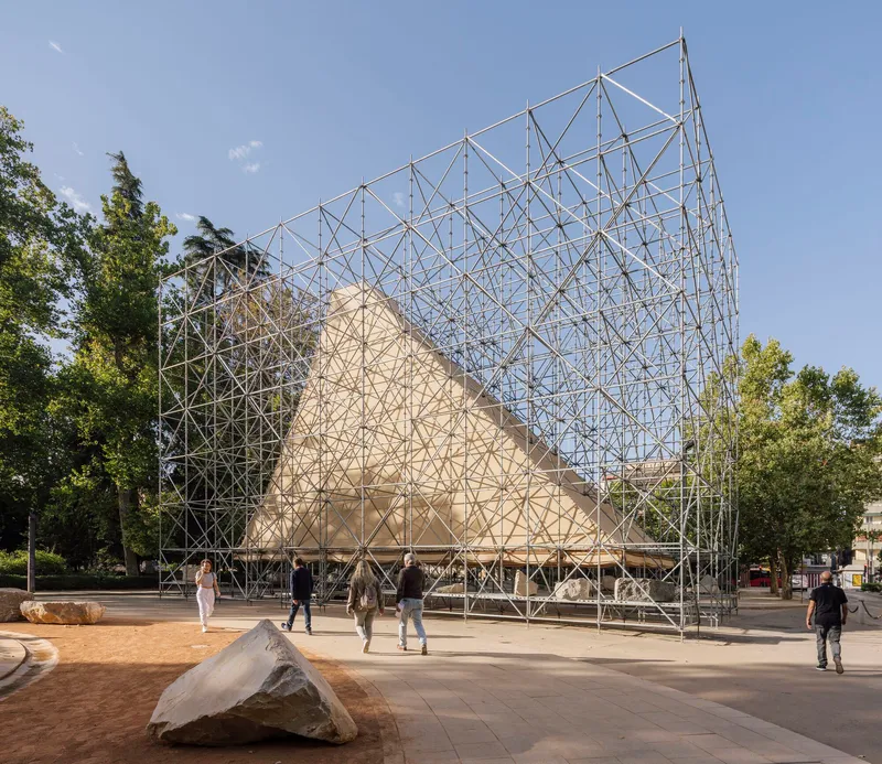 Triangular timber pavilion enclosed by a steel scaffolding framework in a public plaza with scattered stones