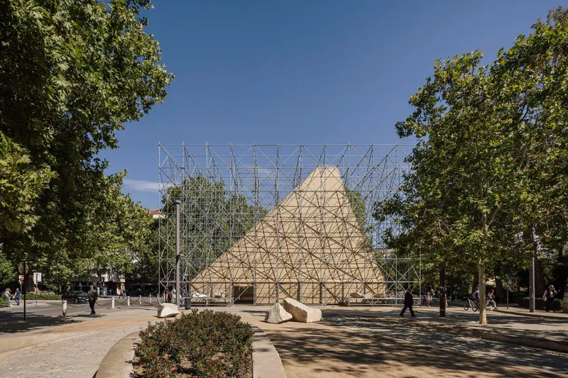 Frontal view of the scaffolding structure framing the timber gable form between tree-lined pathways