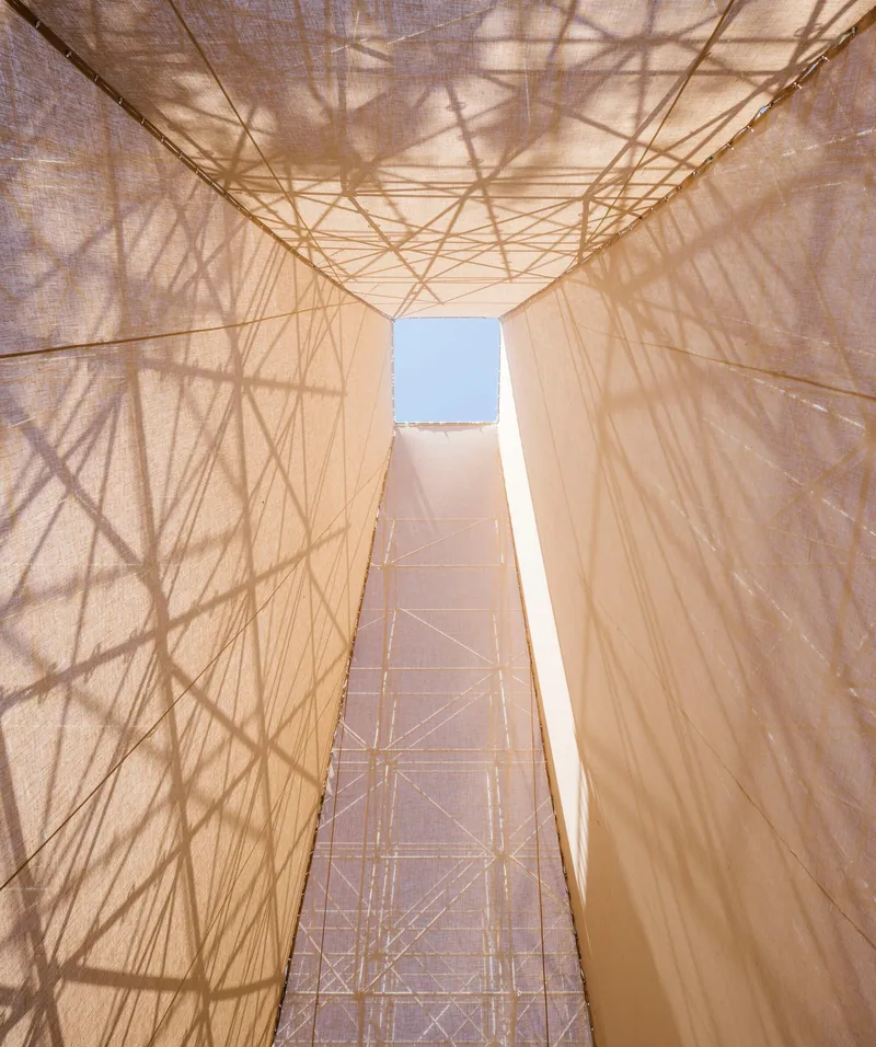 Upward view through the central void showing steel trusses and scaffold shadows on translucent fabric surfaces