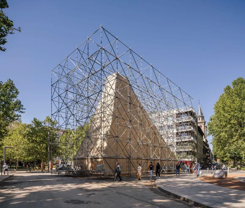 Triangulated steel scaffolding structure enclosing a gabled volume in a tree-lined plaza with pedestrians