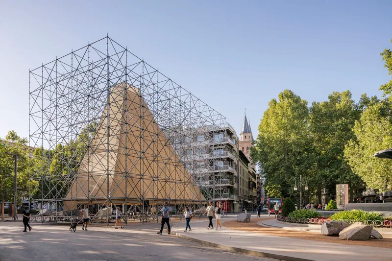 Tubular steel framework defining a house-shaped pavilion with plywood panels on a public square