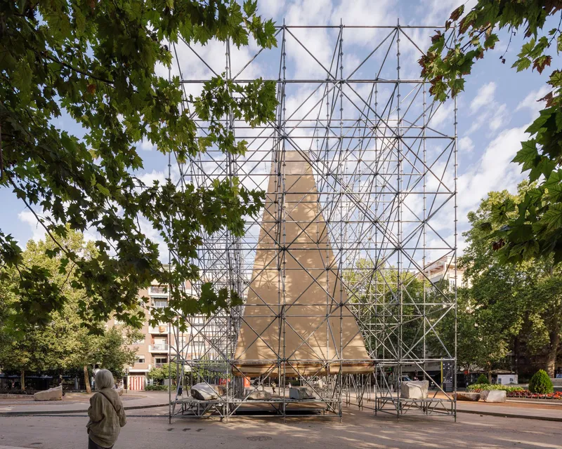 Steel tube scaffolding enclosing a pitched-roof form seen through overhanging tree branches with a visitor nearby