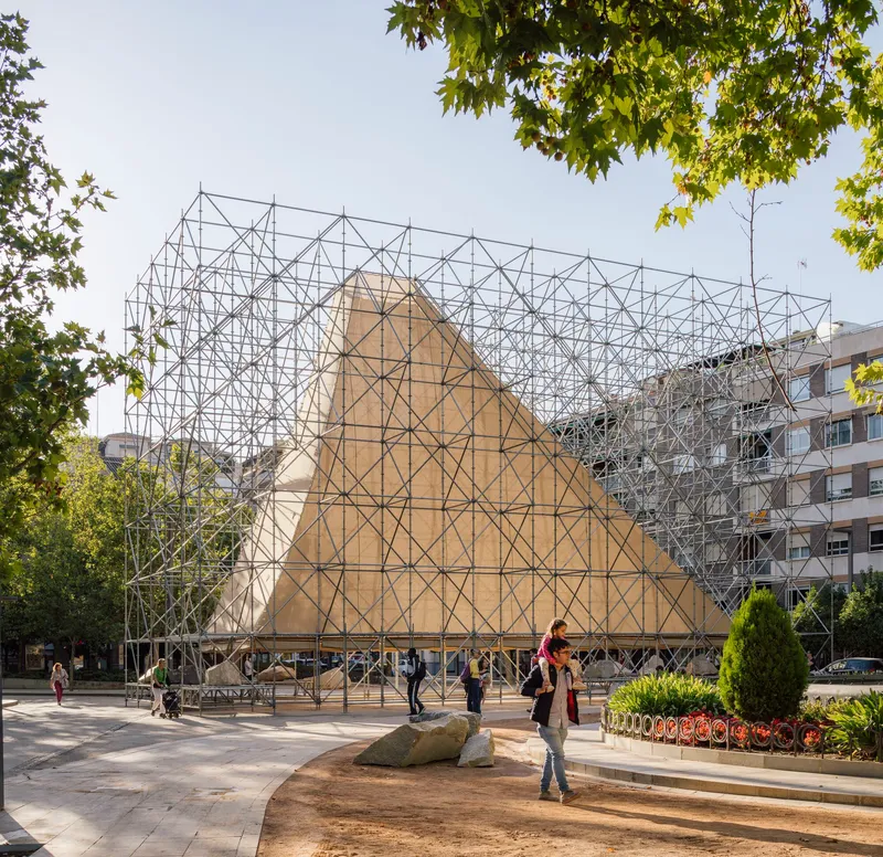 Triangulated metal scaffold pavilion with plywood-clad gable in a plaza with large boulders and planted beds