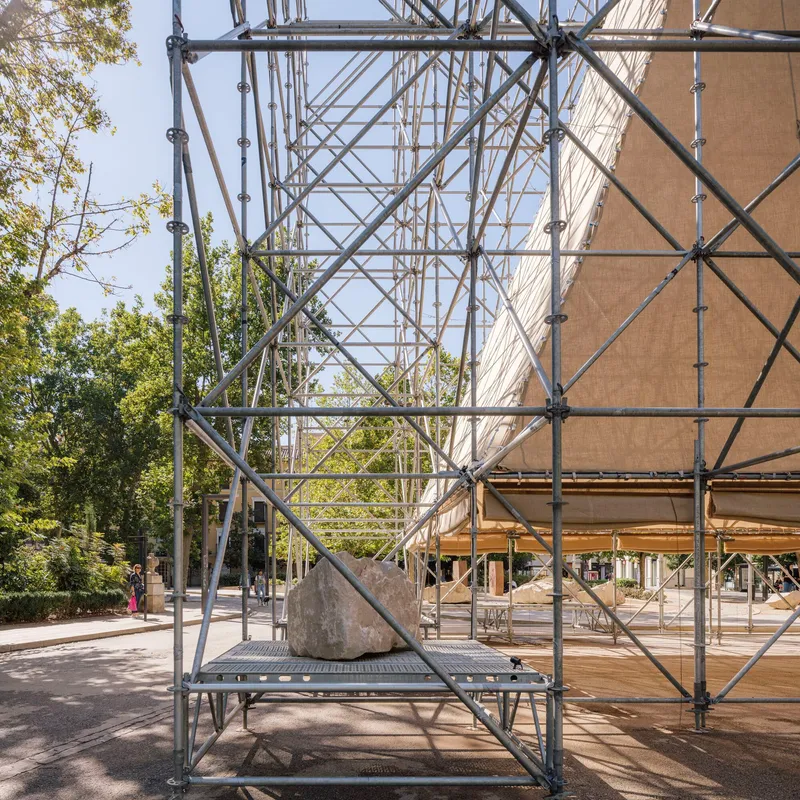 Metal scaffold frame detail with large boulder on platform and tree foliage visible beyond