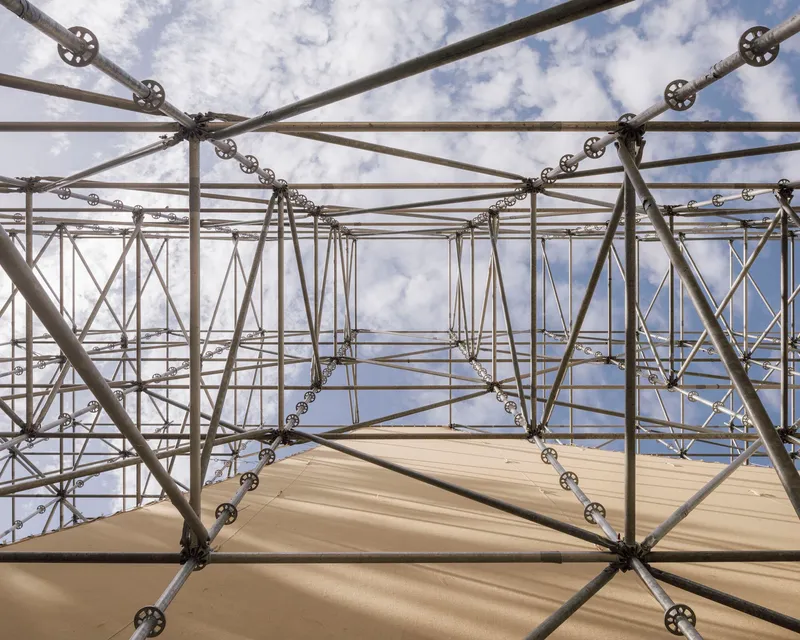 Upward view into scaffolding grid with circular connector nodes and plywood panels against blue sky