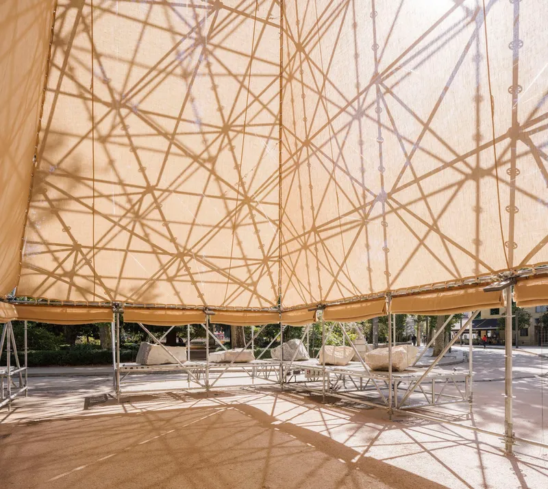 Dappled light patterns from the fabric roof overhead illuminating hammocks suspended beneath the pavilion structure