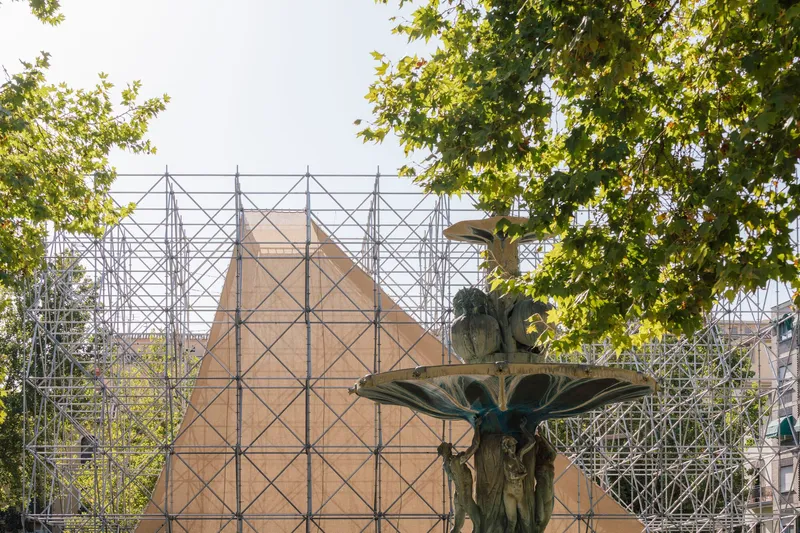 Bronze fountain sculpture framed by scaffolding and fabric screening with dappled sunlight through tree canopy