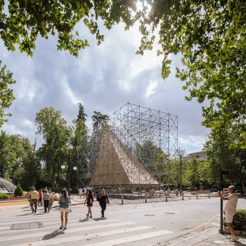 Scaffolding pavilion with burlap panels in a park plaza with visitors and overhanging tree branches