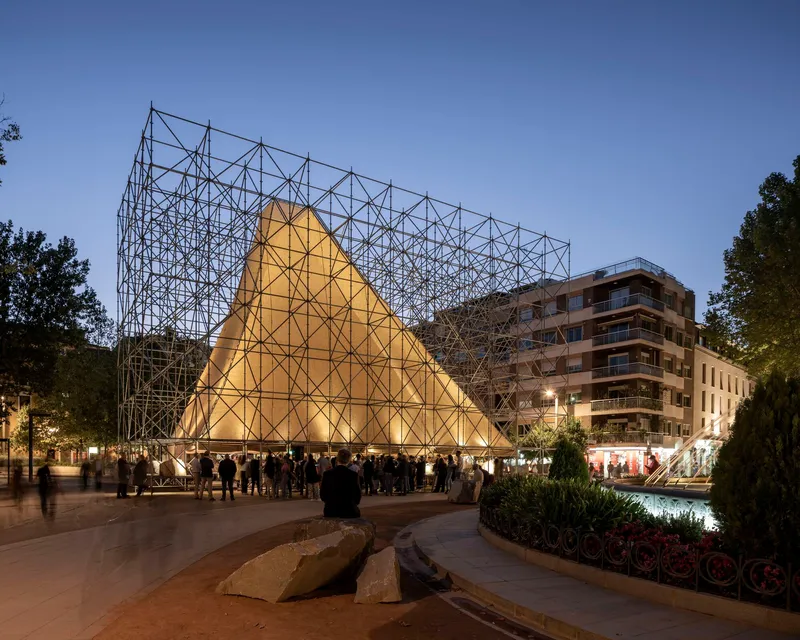 Illuminated fabric-clad scaffolding pyramid in a plaza at dusk with gathered visitors