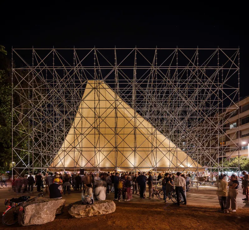 Nighttime view of the glowing scaffold-clad pavilion with crowds gathered around its base