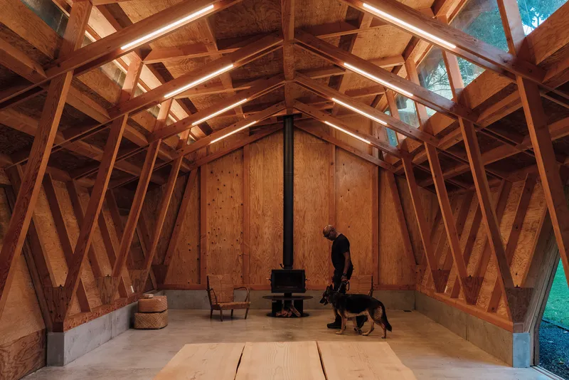 Main hall with a person and dog beside a central stove under the pitched timber frame at dusk