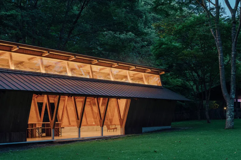 Diagonal braced timber facade with metal standing-seam roof and clerestory band illuminated at twilight