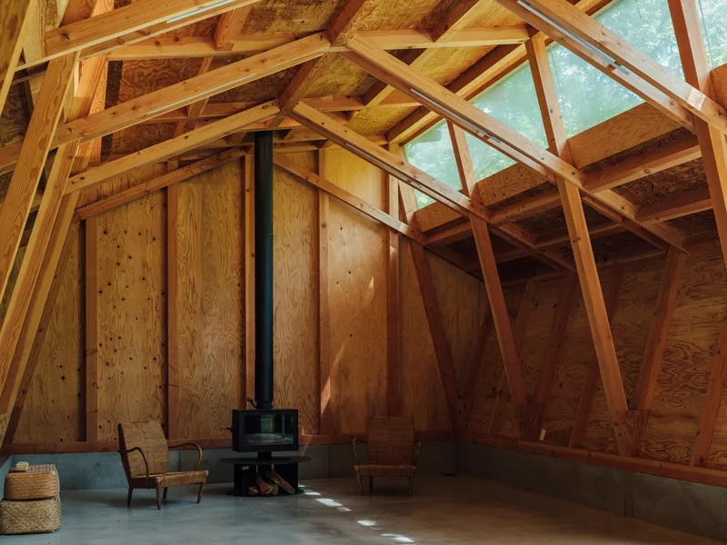 Plywood-clad interior wall with a wood stove on the concrete floor beneath angled timber trusses