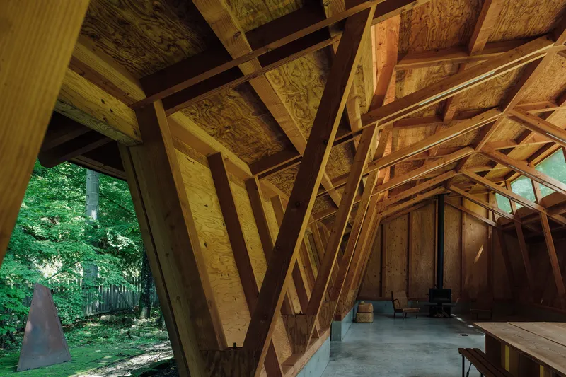 View through diagonal timber portal frames toward the forest and concrete floor in dappled light