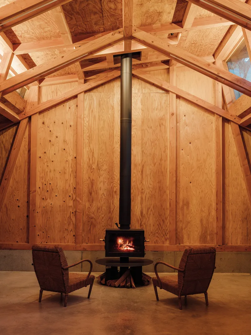 Freestanding wood stove with vertical flue pipe centered between two chairs under the vaulted timber ceiling