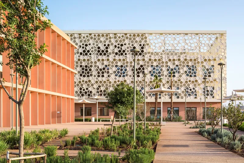 Plaza view showing the perforated honeycomb facade screen with young trees and planting beds in the foreground