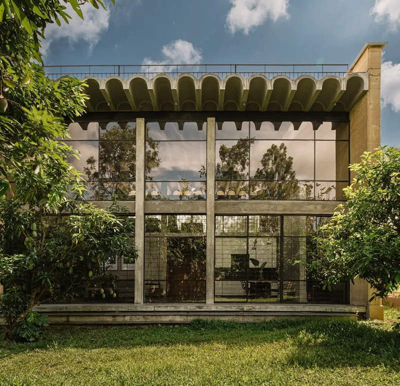 Garden facade with gridded glazing and scalloped concrete canopy framed by lawn and foliage