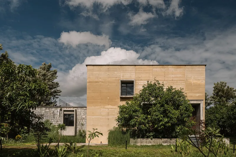 Timber-clad volume with single window rising above a stone base amid tropical vegetation and dramatic clouds