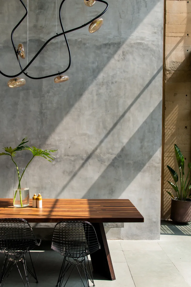 Diagonal shadows falling across concrete wall beside dining table with sculptural pendant light and potted plants