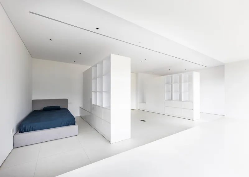 Open-plan bedroom alcove with bed and freestanding white partition wall dividing space from adjacent storage area